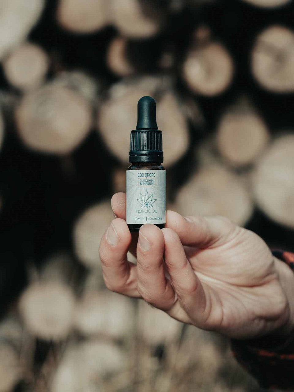 Botella de aceite de CBD Nordic Oil con gotero, sostenida por una mano, fondo desenfocado de troncos.